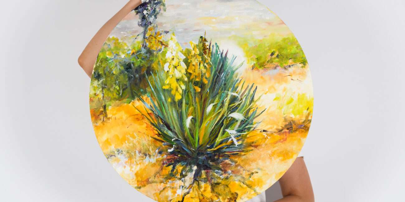Două mâini în mănuși albe țin cu grijă 'Life 3', o pictură circulară vibrantă de Flori Buldus, simbolizând simbioza dintre vegetația mediteraneană și nisipul arid.